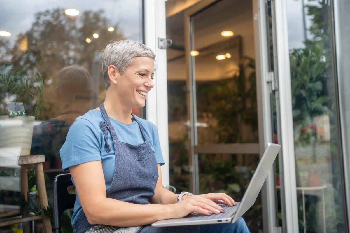 Small flower shop owner florist wearing apron working on laptop in store